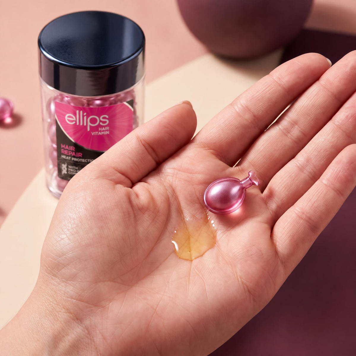 An Ellips Hair Repair capsule twisted open in the palm next to its oil