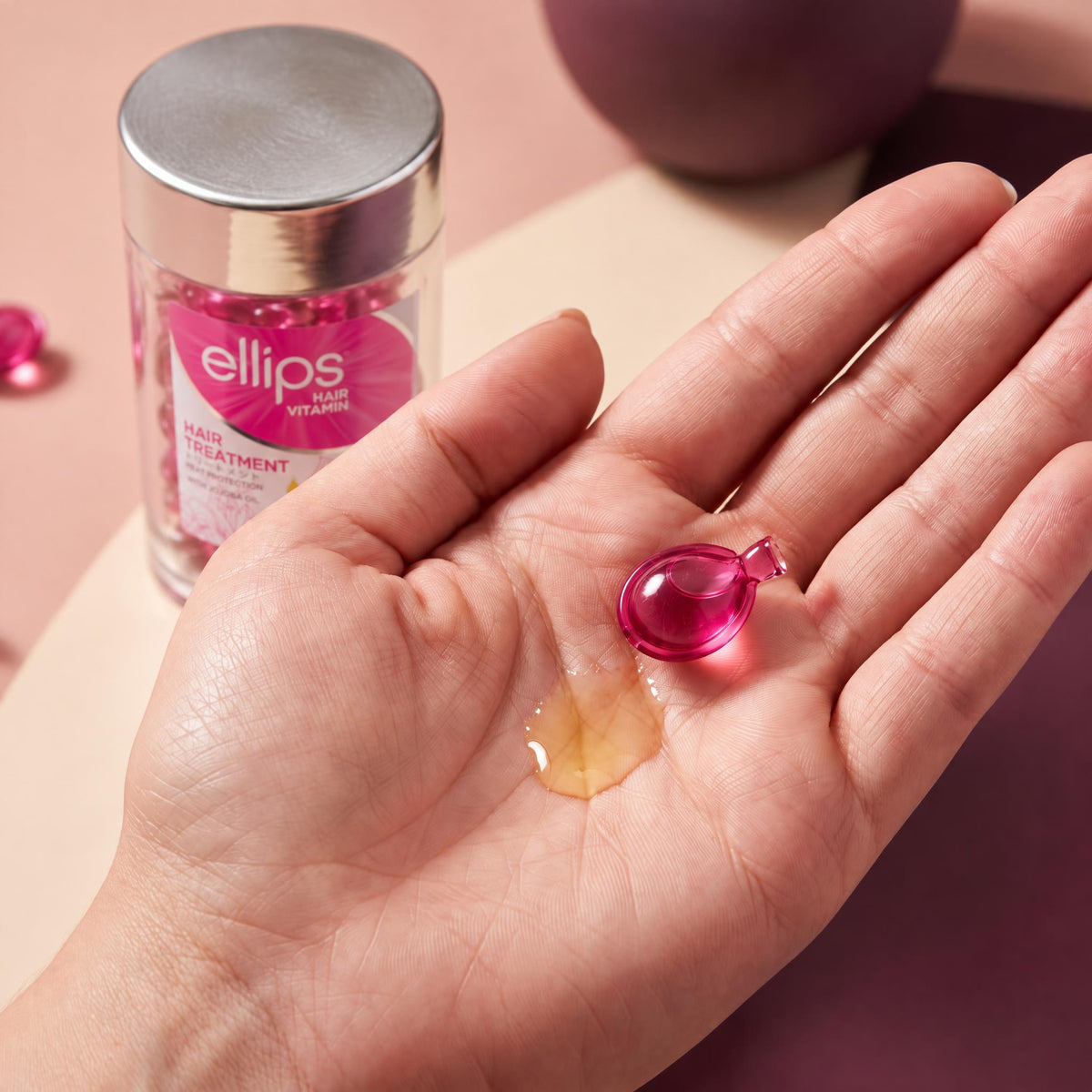 An Ellips Hair Treatment capsule twisted open in the palm next to its oil