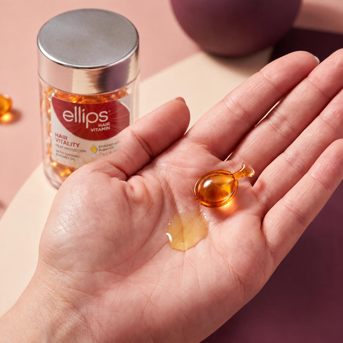 An Ellips Hair Vitality capsule twisted open in the palm next to its oil