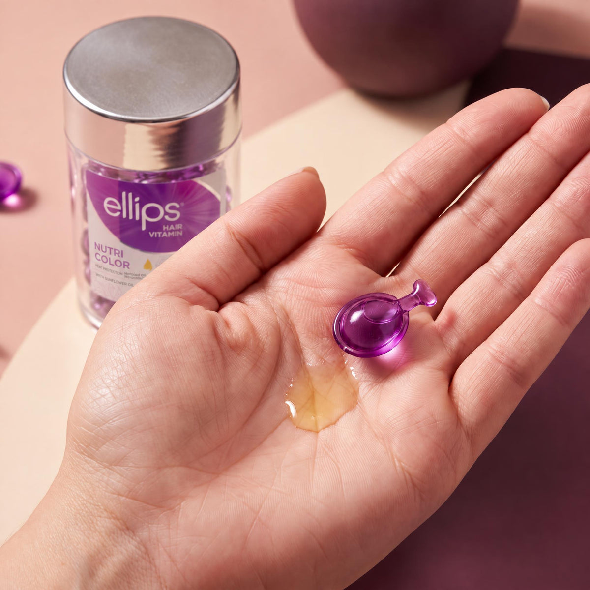 An Ellips Nutri Color capsule twisted open in the palm next to its oil
