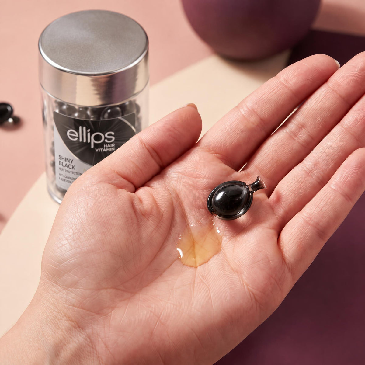 An Ellips Shiny Black capsule twisted open in the palm next to its oil