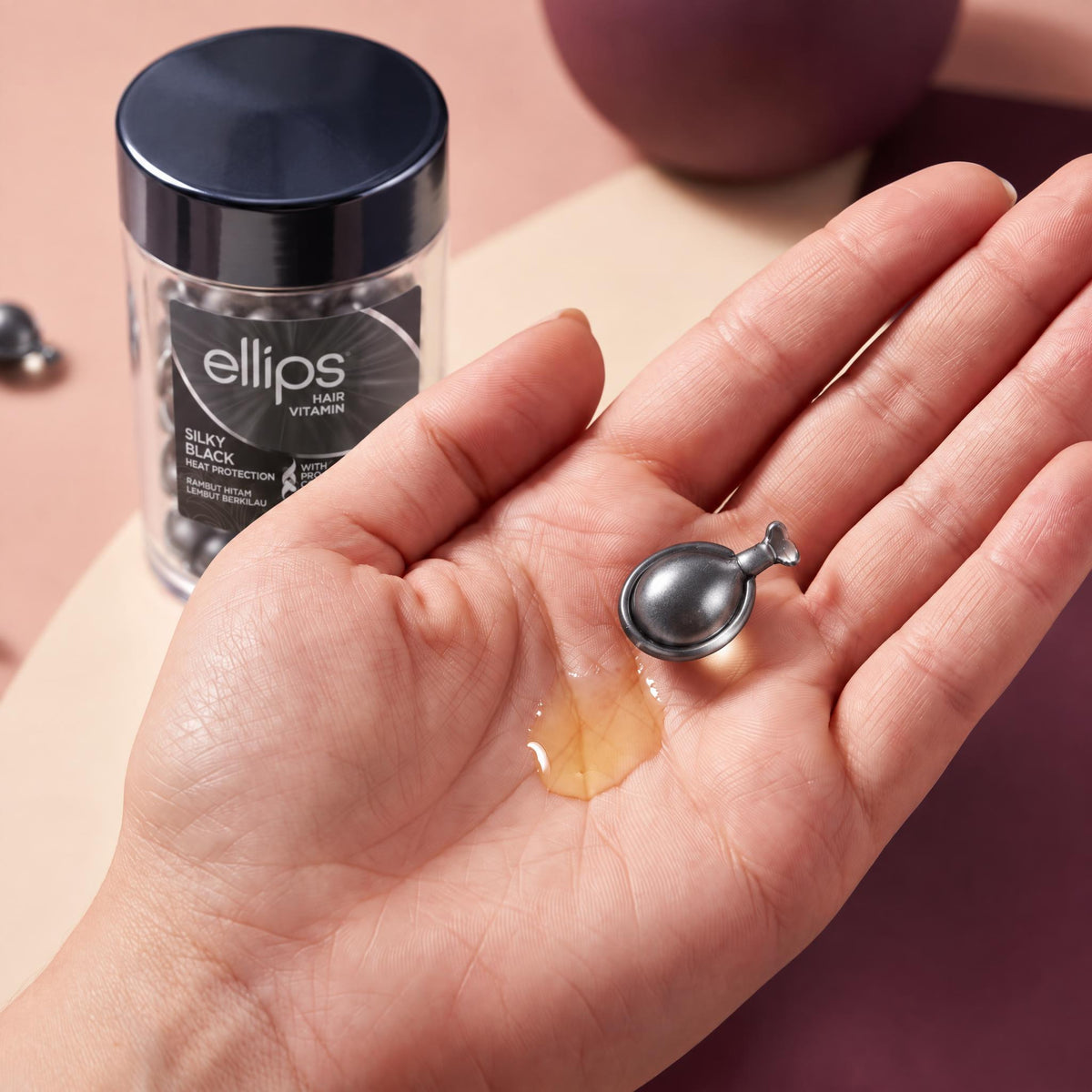 An Ellips Silky Black capsule twisted open in the palm next to its oil