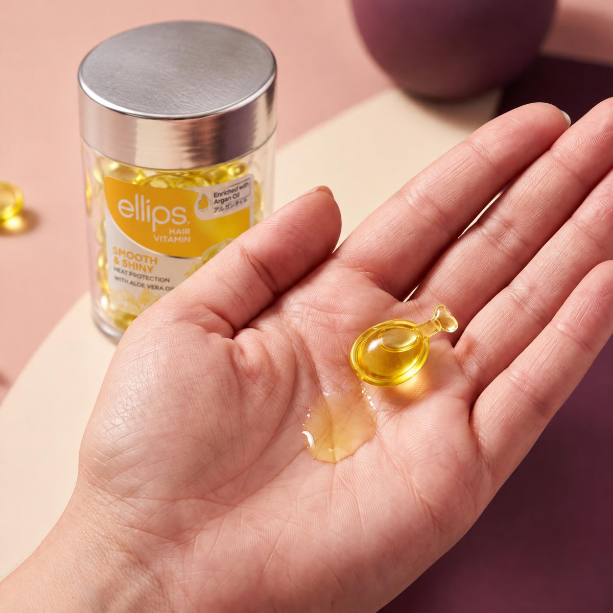 An Ellips Smooth & Shiny capsule twisted open in the palm next to its oil