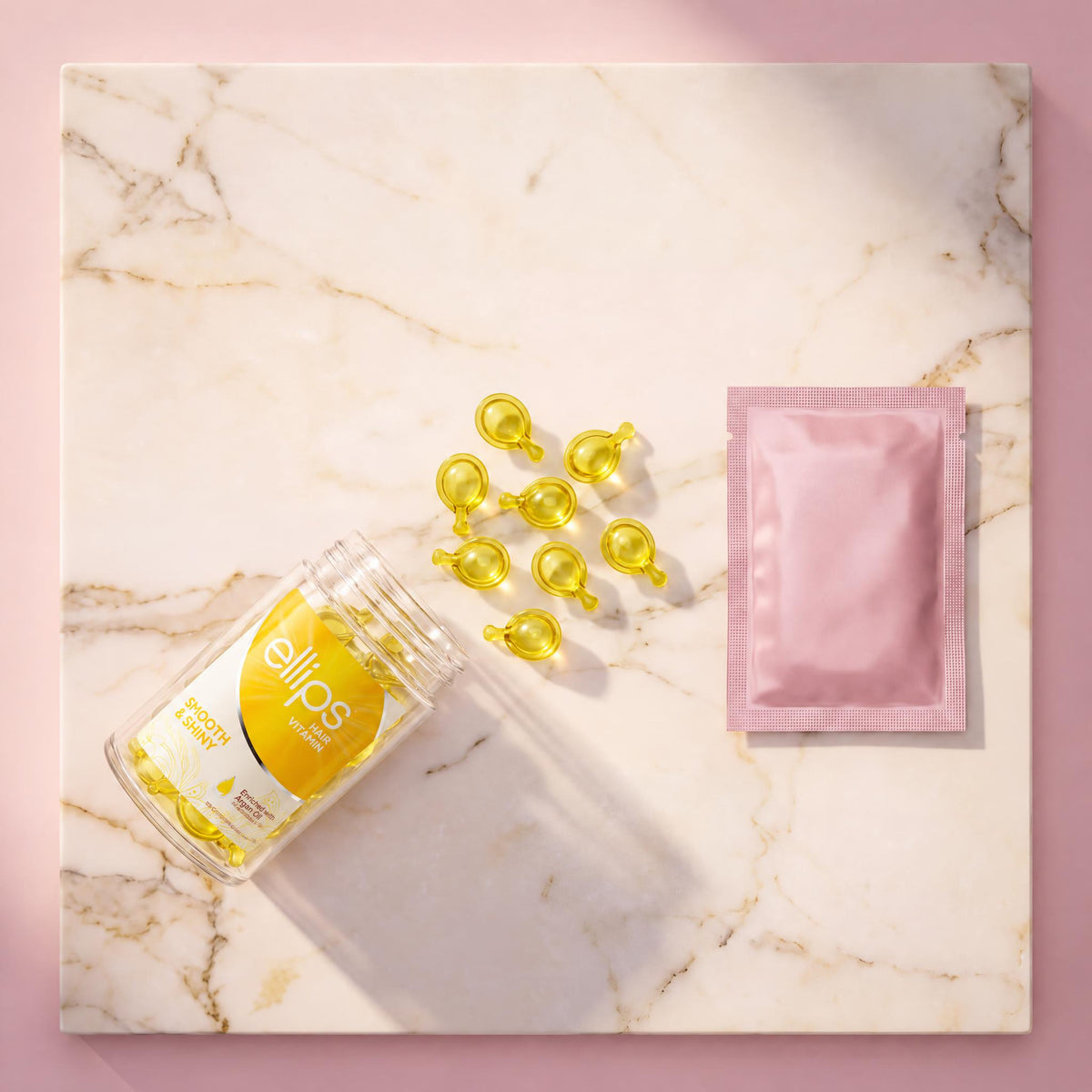 Top view of an open Ellips Smooth & Shiny jar with lemon-yellow capsules and a pink sachet on marble